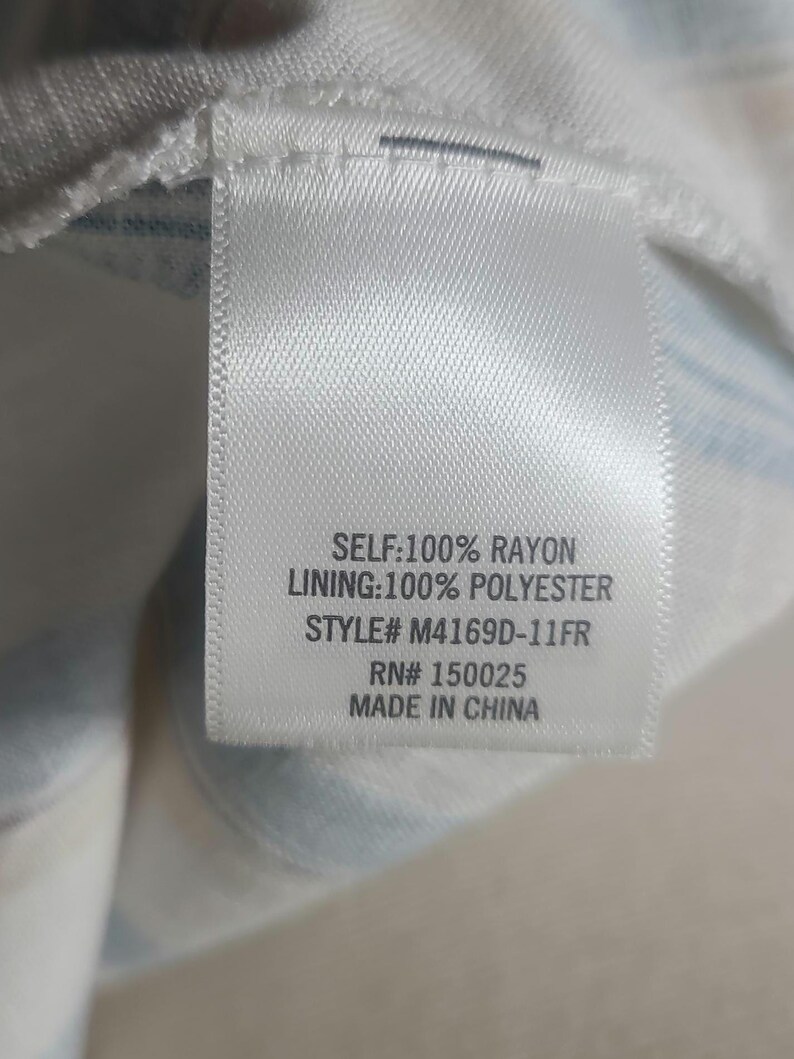 以下が含まれることがあります： 黒い文字が書かれた白い布タグ。タグには「SELF: 100% RAYON, LINING: 100% POLYESTER, STYLE# M4169D-11FR, RN# 150025, MADE IN CHINA」と書かれています。タグは白と青のストライプの生地に付いています。