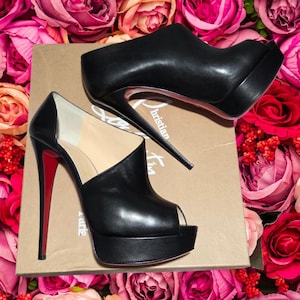 May include: A pair of black leather platform peep-toe heels with red soles. The heels have a stiletto design and are displayed on a cardboard box with the brand name. The background features a vibrant arrangement of pink and red roses.