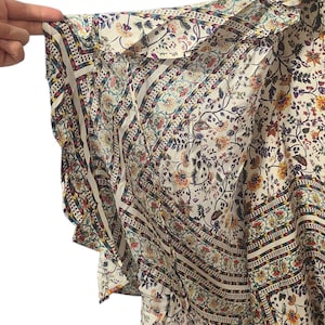 May include: A white floral print top with a V-neck and spaghetti straps. The top features a flowing, ruffled sleeve design with a patterned border in shades of blue, red, and yellow. The fabric is lightweight and airy.