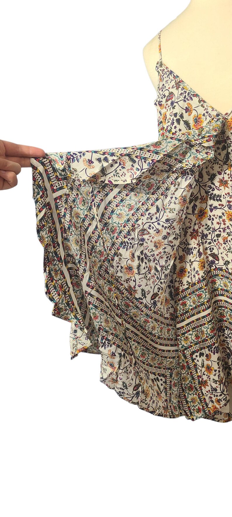 May include: A white floral print top with a V-neck and spaghetti straps. The top features a flowing, ruffled sleeve design with a patterned border in shades of blue, red, and yellow. The fabric is lightweight and airy.