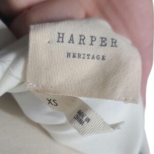 以下が含まれることがあります： 「HARPER HERITAGE」と書かれた濃いグレーの衣類タグのクローズアップ。メインタグの下には「XS」と「MADE IN CHINA」が印刷された小さなタグがあります。生地はオフホワイトです。
