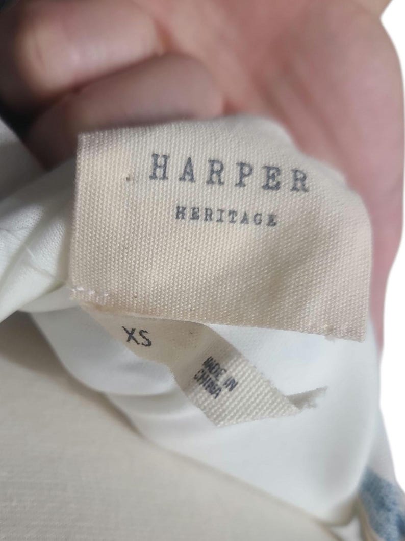 以下が含まれることがあります： 「HARPER HERITAGE」と書かれた濃いグレーの衣類タグのクローズアップ。メインタグの下には「XS」と「MADE IN CHINA」が印刷された小さなタグがあります。生地はオフホワイトです。