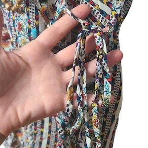 May include: A colorful, patterned tunic top with a floral and geometric design. The top features a mix of red, yellow, and blue flowers on a white background. The sleeves are visible, and the top has tie details.