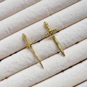 May include: A pair of gold-toned sword earrings. The swords have a detailed design and are set on a white background.