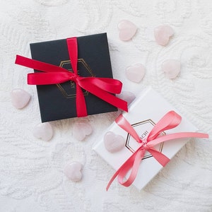 K&ouml;nnte beinhalten: Zwei Geschenkboxen, die mit rosa und rotem Band gebunden sind. Die Boxen befinden sich auf einer wei&szlig;en Oberfl&auml;che mit rosafarbenen herzf&ouml;rmigen Steinen, die um sie herum verstreut sind. Der Text auf den Boxen lautet "Schmuck" und "Sachi".