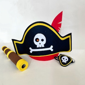 May include: A black felt pirate hat with a yellow trim and a red feather. The hat has a white skull and crossbones design. The set also includes a brown felt telescope and a black felt eye patch with a white skull design.