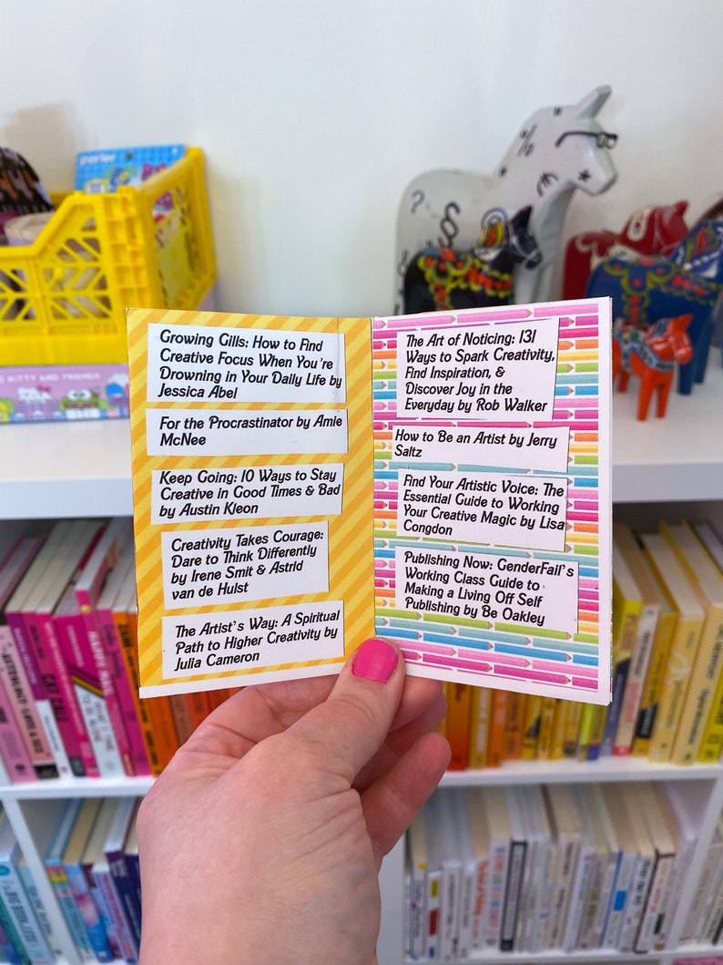 May include: A close-up of a book with a colorful cover and a list of book titles and authors. The book is open to a page with the title "Growing Gills: How to Find Creative Focus When You're Drowning in Your Daily Life" by Jessica Abel. The book is sitting on a shelf with other books.