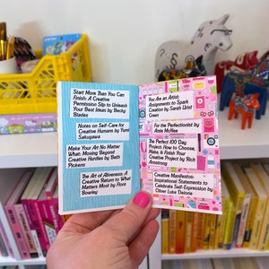 May include: A pink and white book with a variety of colorful stickers on the cover. The book is open to reveal a page with a list of creative books and their authors. The book is being held by a hand with pink nail polish.