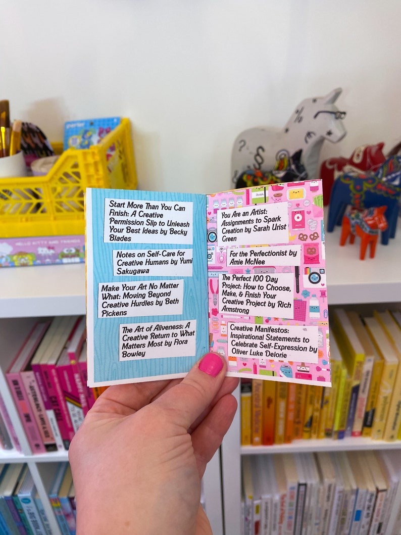 May include: A pink and white book with a variety of colorful stickers on the cover. The book is open to reveal a page with a list of creative books and their authors. The book is being held by a hand with pink nail polish.