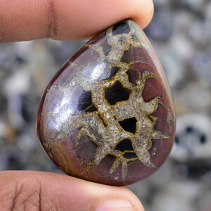 May include: A teardrop-shaped cabochon of red, brown, and black dendritic agate with a silver-colored metallic sheen. The stone has a unique pattern of dendrites, which are tree-like formations of minerals.