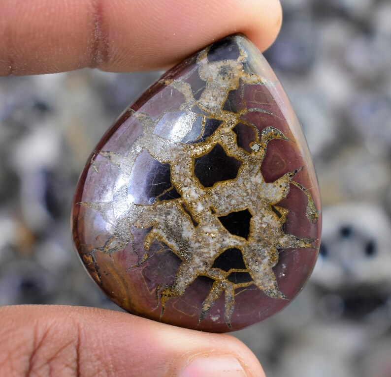 May include: A teardrop-shaped cabochon of red, brown, and black dendritic agate with a silver-colored metallic sheen. The stone has a unique pattern of dendrites, which are tree-like formations of minerals.