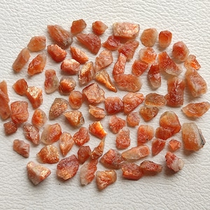 May include: A collection of raw, orange-brown sunstone crystals. The crystals are various sizes and shapes, with some having a shimmery, iridescent quality.