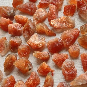 May include: A collection of raw, orange-brown sunstone crystals. The crystals are rough and have a natural, unpolished finish. They are scattered on a white surface.