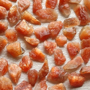 May include: A collection of raw, orange-brown sunstone crystals. The crystals are various sizes and shapes, with some having a shimmery, iridescent quality.