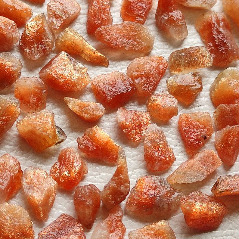 May include: A collection of raw, orange-brown sunstone crystals. The crystals are various sizes and shapes, with some having a shimmery, iridescent quality.