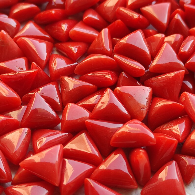 May include: A close-up view of a pile of triangular red coral gemstones. The coral is polished and has a smooth, glossy finish.