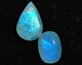 May include: Two iridescent moonstone cabochons, one teardrop shaped and one oval, with a blue and white sheen on a black background.