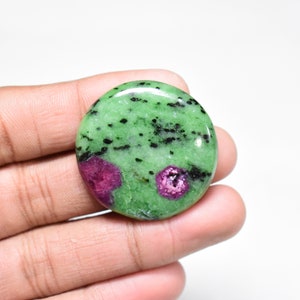 May include: A round, green and pink zoisite cabochon gemstone with black inclusions. The stone is polished and has a smooth surface.