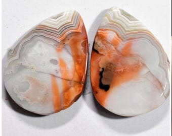 May include: A pair of agate cabochons with a white, orange, and brown color pattern. The cabochons are teardrop shaped and have a smooth, polished surface.