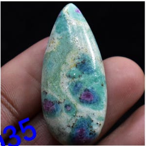 May include: A teardrop-shaped cabochon gemstone with a green, blue, and pink colour pattern. The gemstone has a smooth, polished surface.