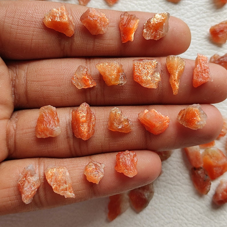 May include: A collection of raw, orange sunstone crystals. The crystals are various sizes and shapes, with a shimmery, iridescent quality.