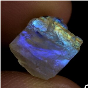 Puede incluir: Una gema bruta e iridiscente con matices azules, verdes y amarillos. La piedra es translúcida y tiene una superficie lisa y pulida.