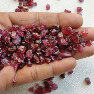 Puede incluir: Un puñado de piedras preciosas de color rojo oscuro en bruto. Se aprecian las formas irregulares de las piedras, con tamaños variables y una calidad ligeramente translúcida. Las gemas están dispersas por la palma, con algunos trozos sueltos alrededor de la mano.