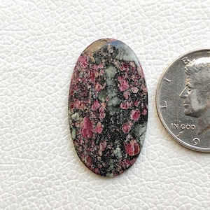 May include: Oval cabochon gemstone with a black, green, and pink speckled pattern. The stone is approximately the size of a US quarter.