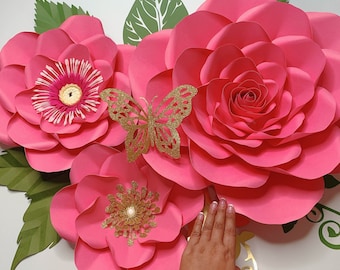 May include: Three large pink paper flowers with a gold butterfly in the center. The flowers are layered and have a ruffled appearance. The flowers are on a white background.