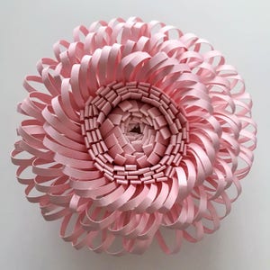K&ouml;nnte beinhalten: Eine rosa Papierblume mit einem geschichteten, spiralf&ouml;rmigen Design. Die Blume besteht aus vielen d&uuml;nnen Papierstreifen, die gefaltet und &uuml;bereinander gelegt werden, um einen dreidimensionalen Effekt zu erzielen.