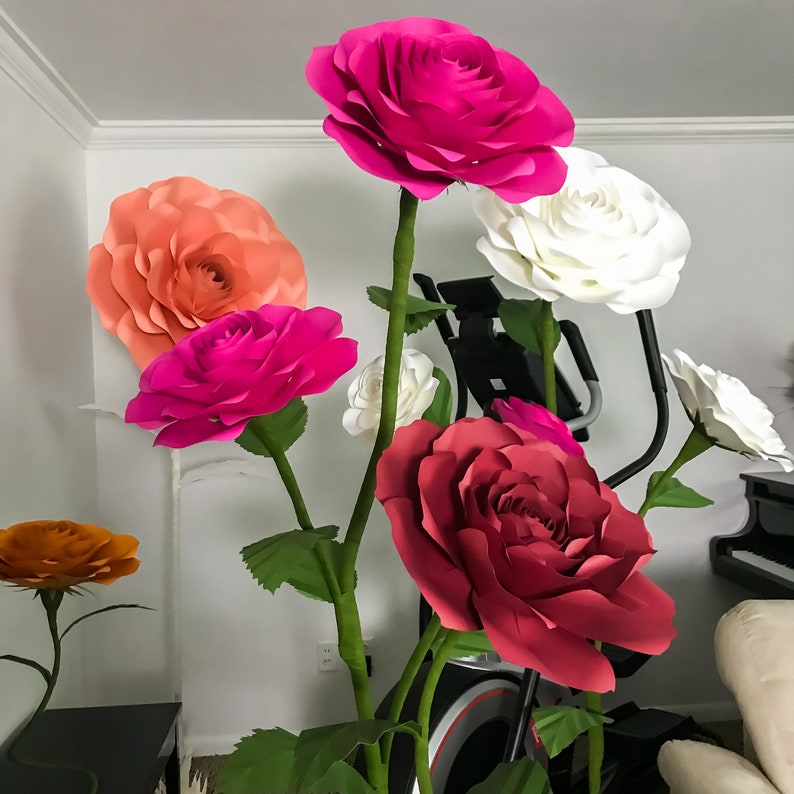 May include: A bouquet of large paper flowers in various colors, including pink, red, white, and orange. The flowers are arranged in a vase and are displayed in a room with a white wall and a black exercise machine in the background.