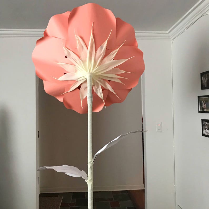 May include: A large, pink paper flower with white petals in the center. The flower is attached to a white stem with green paper leaves.