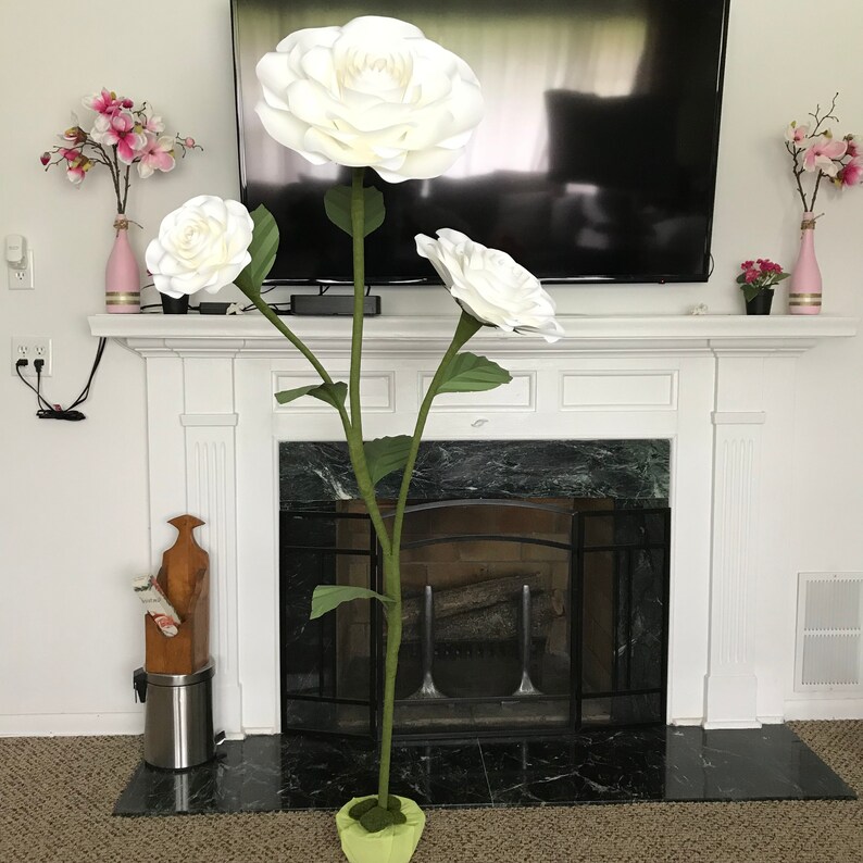 May include: A large white paper flower arrangement with three flowers and green stems. The flowers are arranged in a vase in front of a fireplace.