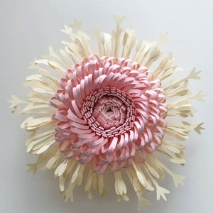 Puede incluir: Una gran escultura de flores de papel con un dise&ntilde;o en capas. El centro es una espiral de papel rosa, rodeada de anillos conc&eacute;ntricos de papel rosa. Los p&eacute;talos exteriores son de color crema y puntiagudos, creando un efecto de explosi&oacute;n estelar. La obra est&aacute; montada sobre un fondo blanco.