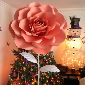 May include: A large, peach-colored paper rose with white paper leaves is attached to a white pole. The rose is in front of a decorated Christmas tree with colorful ornaments and lights.