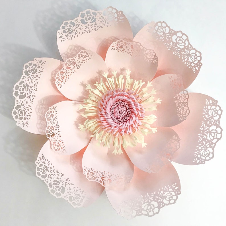 Puede incluir: Una gran flor de papel rosa claro con intrincados cortes tipo encaje en los bordes de los p&eacute;talos. El centro de la flor presenta un dise&ntilde;o en capas de papel rosa y crema, creando una apariencia detallada y texturizada. Es un art&iacute;culo decorativo.
