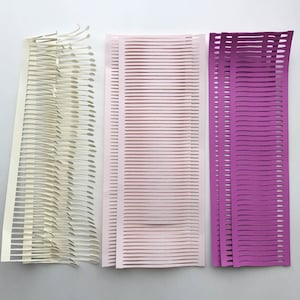 Puede incluir: Tres piezas de arte abstracto en papel en crema, rosa claro y morado. La pieza color crema tiene un dise&ntilde;o &uacute;nico en capas con bordes curvos. Las piezas rosa y morada presentan l&iacute;neas de corte horizontales, creando un efecto texturizado.