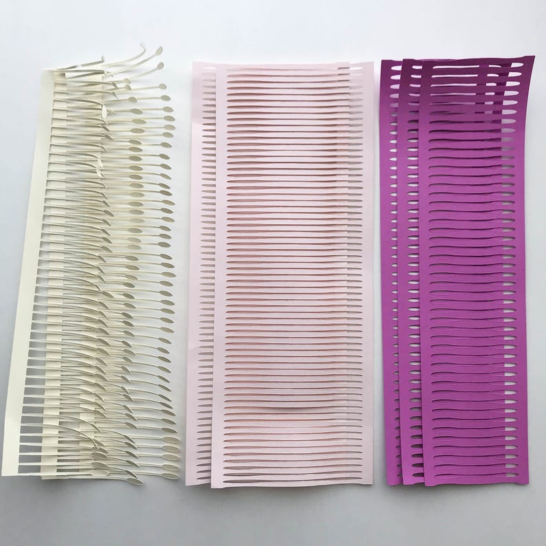 Puede incluir: Tres piezas de arte abstracto en papel en crema, rosa claro y morado. La pieza color crema tiene un dise&ntilde;o &uacute;nico en capas con bordes curvos. Las piezas rosa y morada presentan l&iacute;neas de corte horizontales, creando un efecto texturizado.