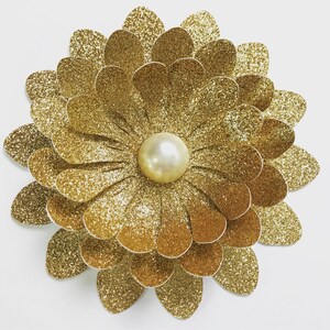 Puede incluir: Una gran flor de brillantina dorada con una perla en el centro. La flor está hecha de varias capas de papel.