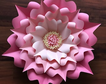 May include: A large pink paper flower with multiple layers of petals. The center of the flower is a smaller white flower with a yellow center.