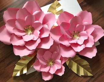 May include: Three large pink paper flowers with gold centers and two gold paper leaves. The flowers are layered and have a delicate, ruffled texture.