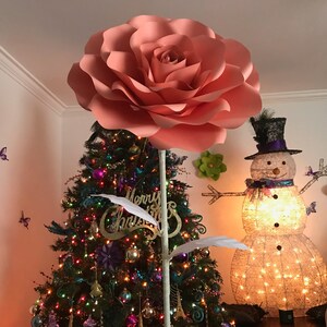 May include: A large, peach-colored paper flower with a white stem stands in front of a decorated Christmas tree. The tree is decorated with colorful ornaments, lights, and a "Merry Christmas" sign. A snowman made of lights stands to the right of the flower.