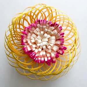 Puede incluir: Una flor de papel amarilla con un centro rosa y blanco. La flor está hecha de muchas capas de papel que están enroscadas y retorcidas para crear un efecto 3D.