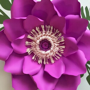 Puede incluir: Una gran flor de papel magenta vibrante con un centro detallado. Los p&eacute;talos de la flor est&aacute;n en capas, creando profundidad y textura. El centro presenta un dise&ntilde;o rosa claro y crema. Se ven hojas verdes en el fondo.