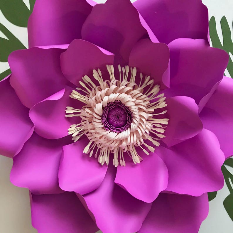 Puede incluir: Una gran flor de papel magenta vibrante con un centro detallado. Los p&eacute;talos de la flor est&aacute;n en capas, creando profundidad y textura. El centro presenta un dise&ntilde;o rosa claro y crema. Se ven hojas verdes en el fondo.