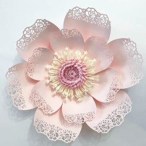 K&ouml;nnte beinhalten: Eine gro&szlig;e, rosafarbene Papierblume mit filigranen Spitzendetails und einem gelben und rosafarbenen Zentrum. Die Blume besteht aus mehreren Papierschichten und hat ein zartes, feminines Aussehen.