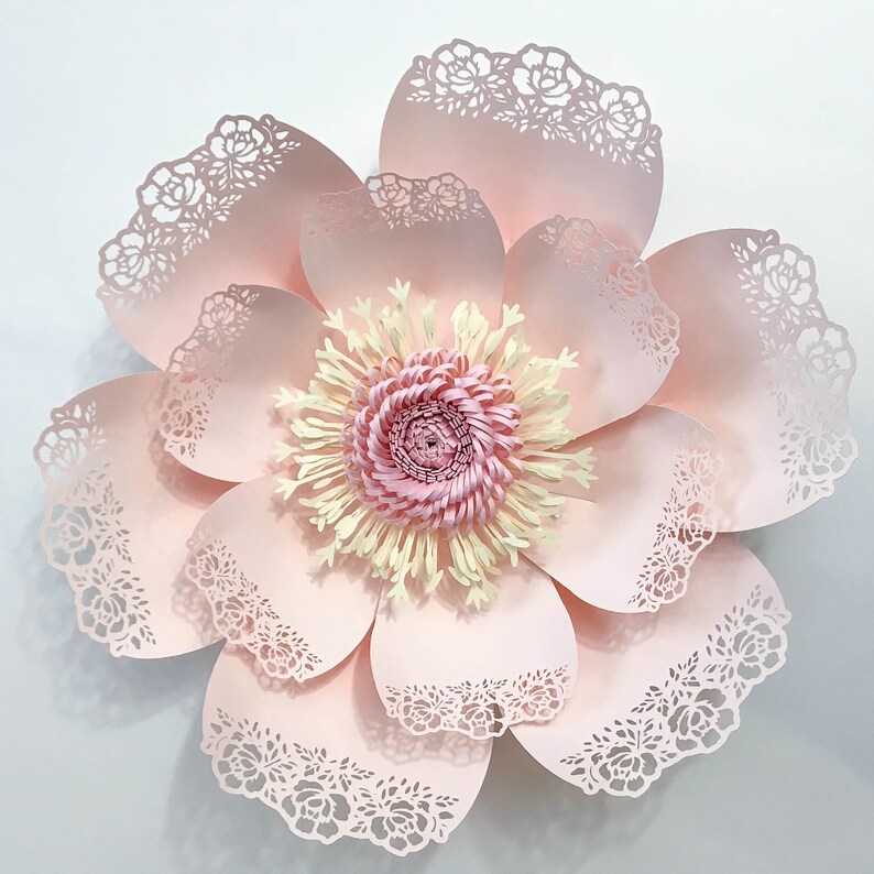 K&ouml;nnte beinhalten: Eine gro&szlig;e, rosafarbene Papierblume mit filigranen Spitzendetails und einem gelben und rosafarbenen Zentrum. Die Blume besteht aus mehreren Papierschichten und hat ein zartes, feminines Aussehen.
