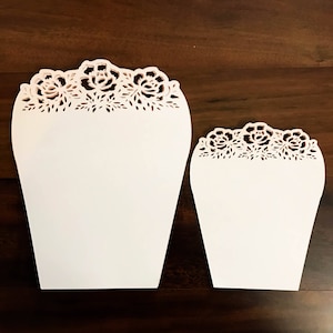 K&ouml;nnte beinhalten: Zwei wei&szlig;e Papierkegel mit einem zarten floralen Spitzenmuster. Die Kegel sind unterschiedlich gro&szlig;, wobei der gr&ouml;&szlig;ere Kegel h&ouml;her ist als der kleinere.