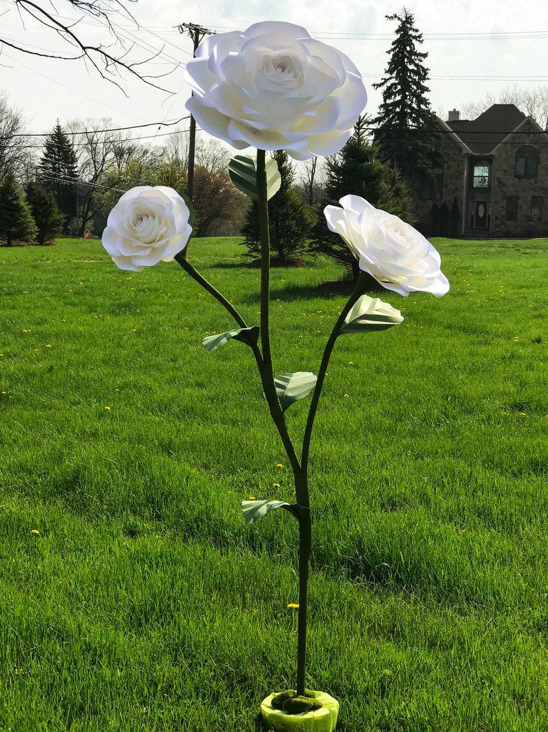 May include: A white paper flower arrangement with three large flowers on a green stem. The flowers are arranged in a staggered pattern, with the largest flower at the top. The flowers are in a grassy field with a house in the background.
