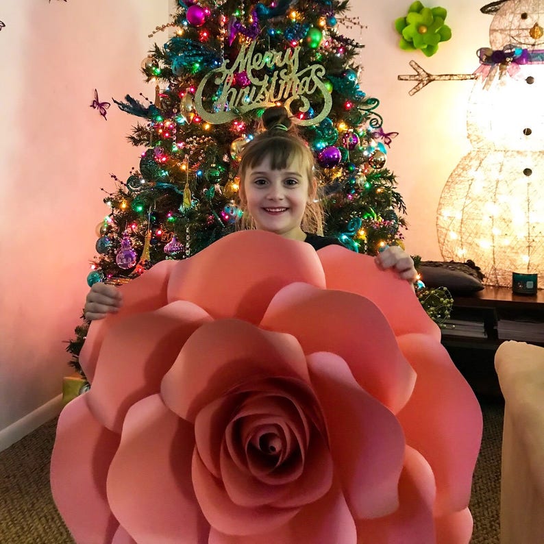 May include: A young girl smiles while holding a large, pink paper flower in front of a decorated Christmas tree. The flower is made of multiple layers of paper and has a large, central rose.